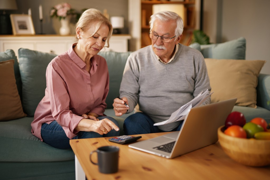 Senior couple reviewing life insurance policy documents and finances on a laptop and with a calculator at home.