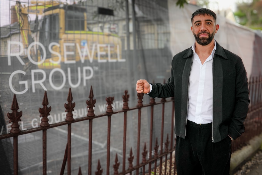 A dark-haired, bearded man stands next to a construction site.
