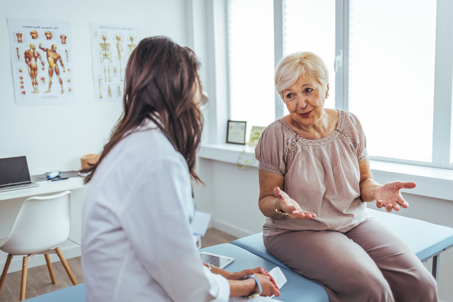 A senior woman on an exam table is having a conversation with a doctor in a modern medical clinic, conveying trust and health-focused interaction. The room includes anatomical charts a