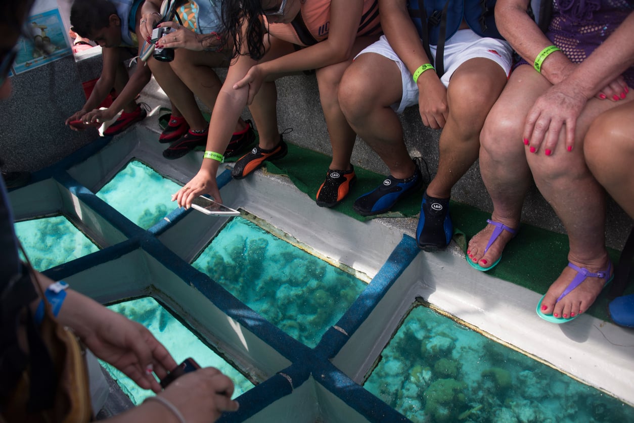 A group of people with colorful wristbands sit on a boat with glass-bottom panels, looking down at the clear water and coral reefs below—one of the region’s most popular tourist attractions. Some are taking photos or reaching toward the glass.