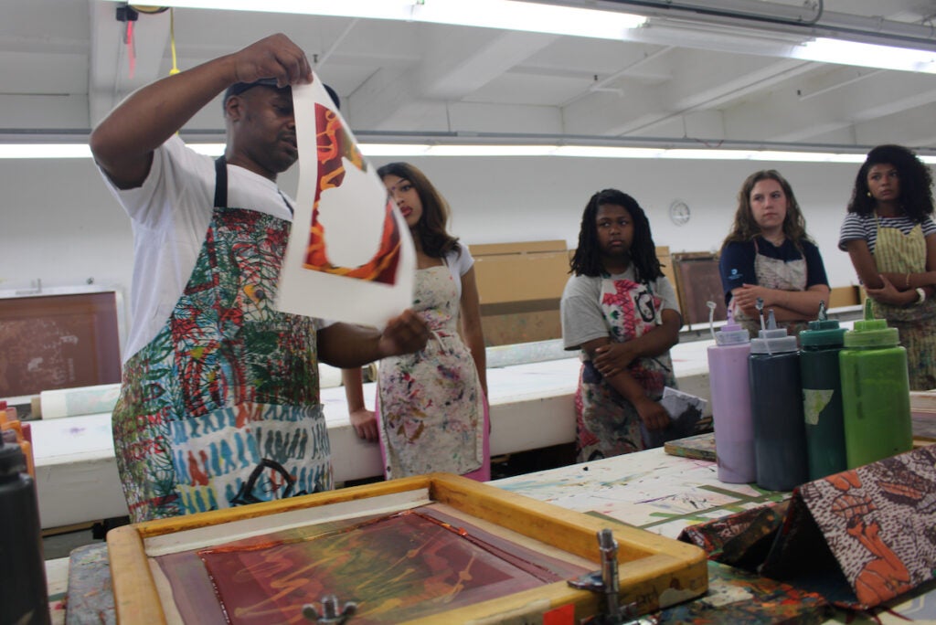 A man holds up an unfinished painting in a workshop in front of four women