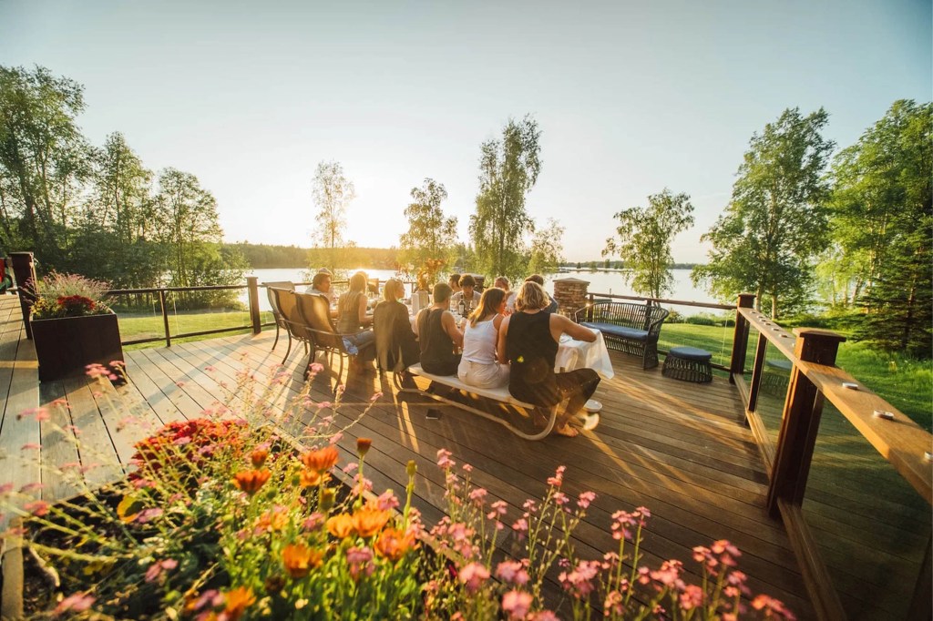 Family and friends gathered for dinner on a wooden deck overlooking a lake at sunset.