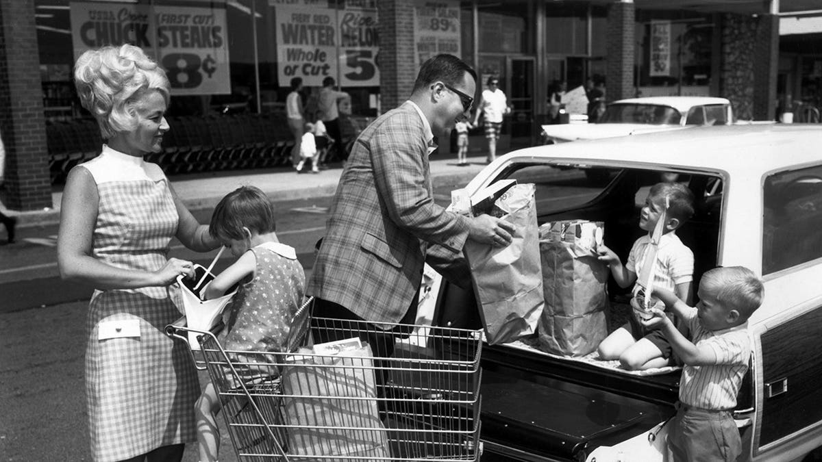 Man loads groceries into a station wagon as two boys help and a girl sits in a shopping cart.