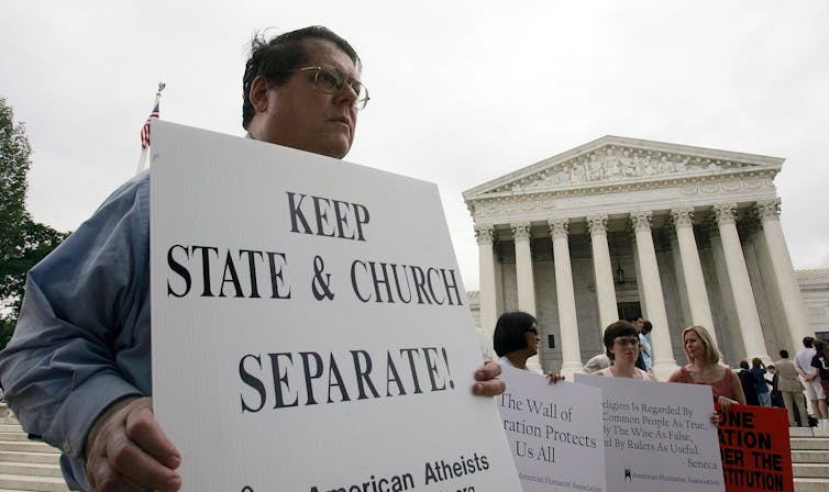 Several protestors stand in front of the U.S. Supreme Court building holding signs, one of which says, 'Keep State & Church Separate.'