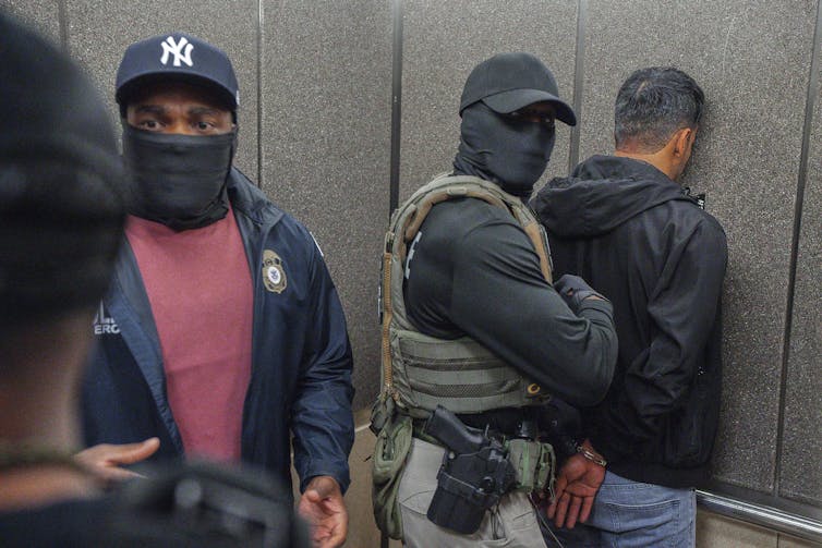 An ICE agent arrests a person with their face against a wall while another is speaking.
