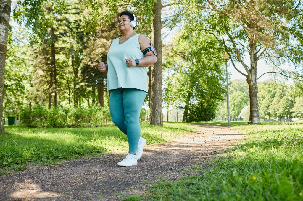 A smiling senior Black woman jogs through a park, wearing headphones and an armband for her phone.