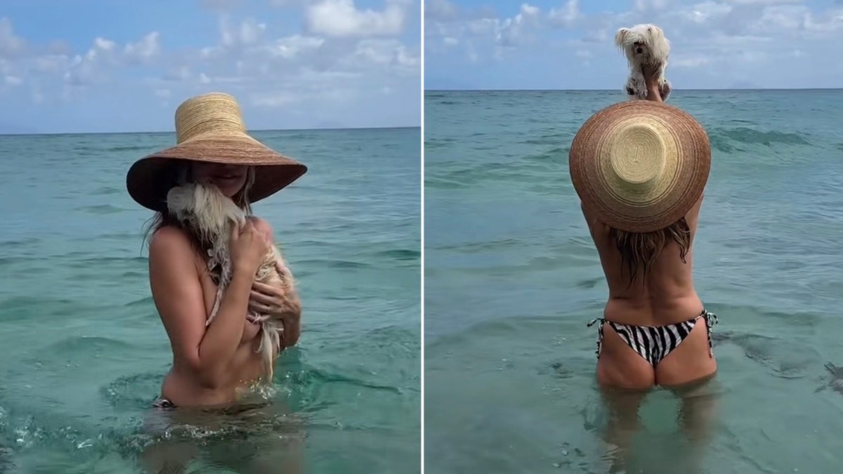 Heidi Klum stands waist-deep in the ocean and lifts a small white dog above her head while wearing a wide-brimmed straw hat.