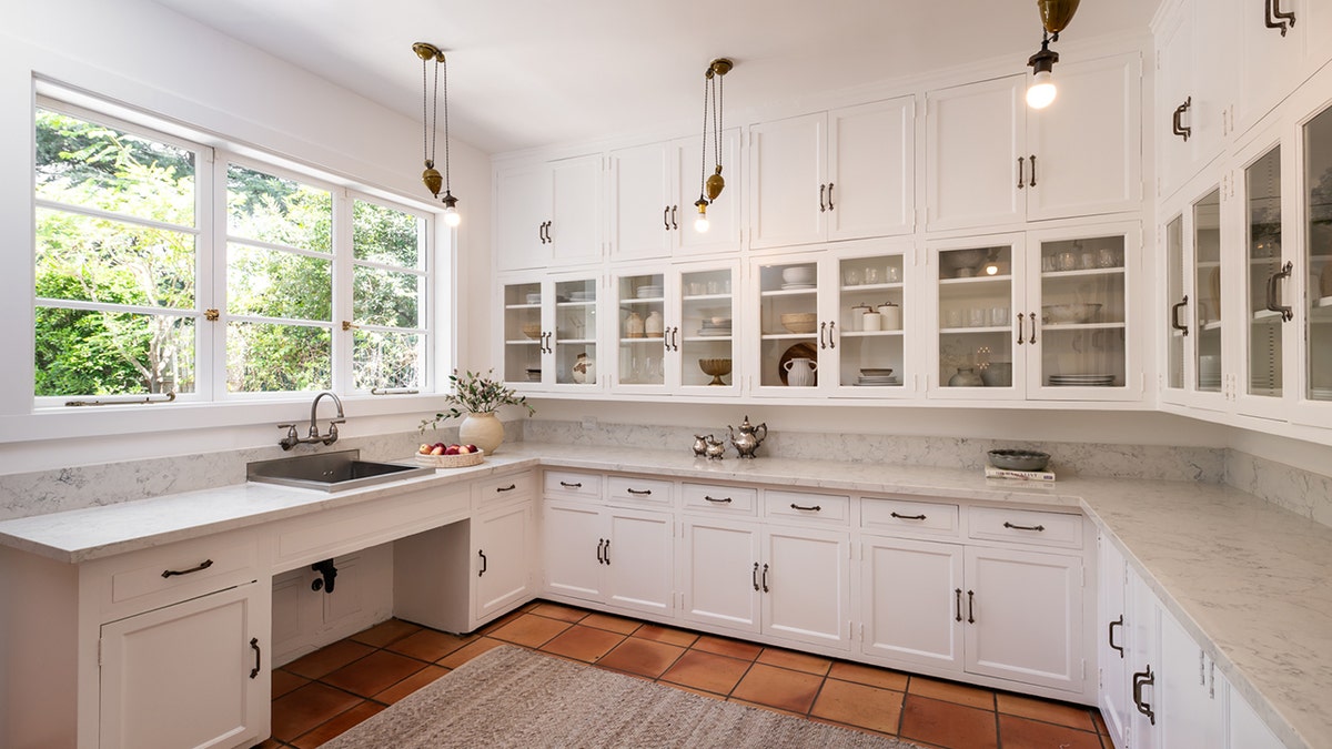 A white kitchen features marble countertops, glass-front cabinets and large windows.