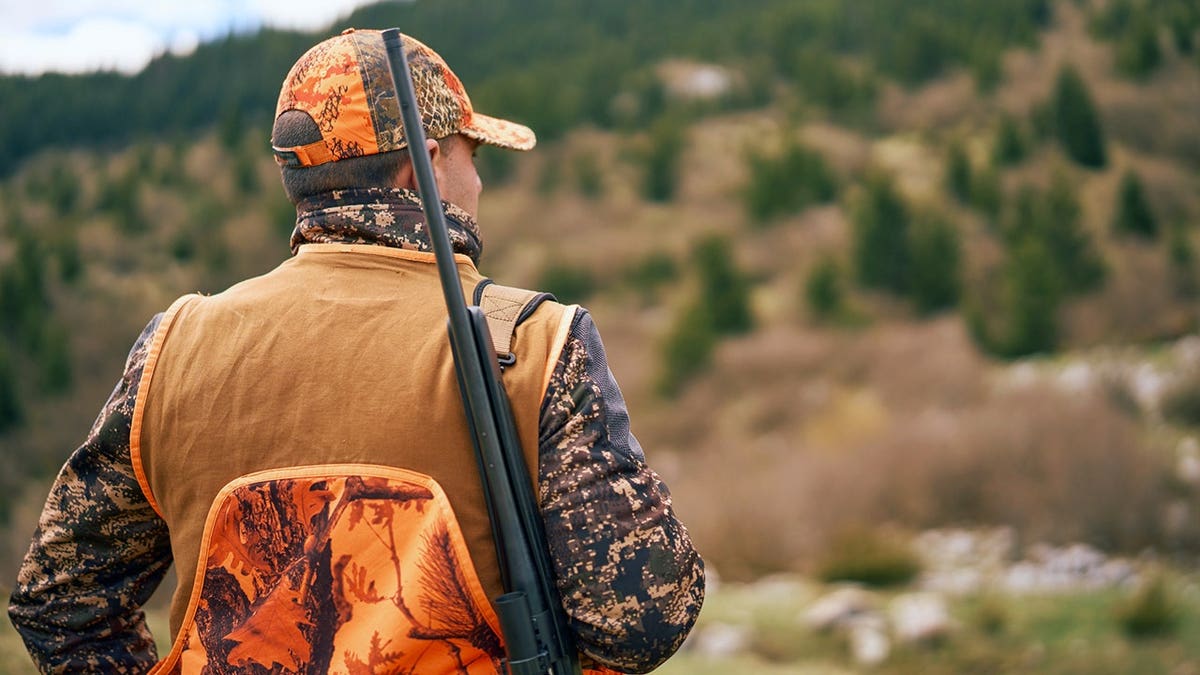 Hunter in blaze orange and camouflage stands in a mountainous forest carrying a rifle.