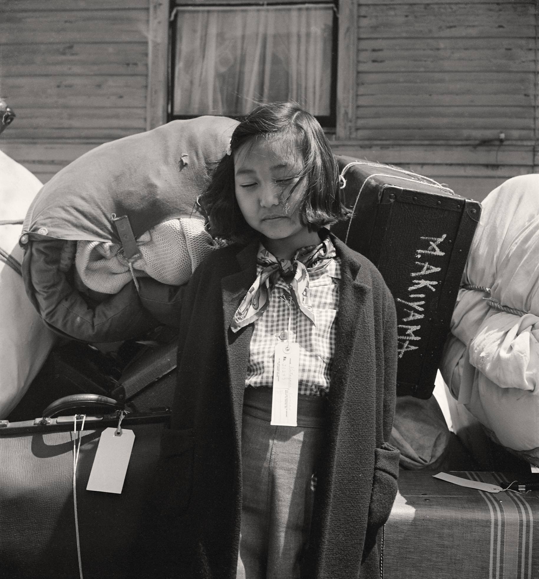 A young Japanese American girl with an identification tag pinned to her coat stands exhausted among piled luggage and bundled belongings during the 1942 wartime evacuation of the West Coast.