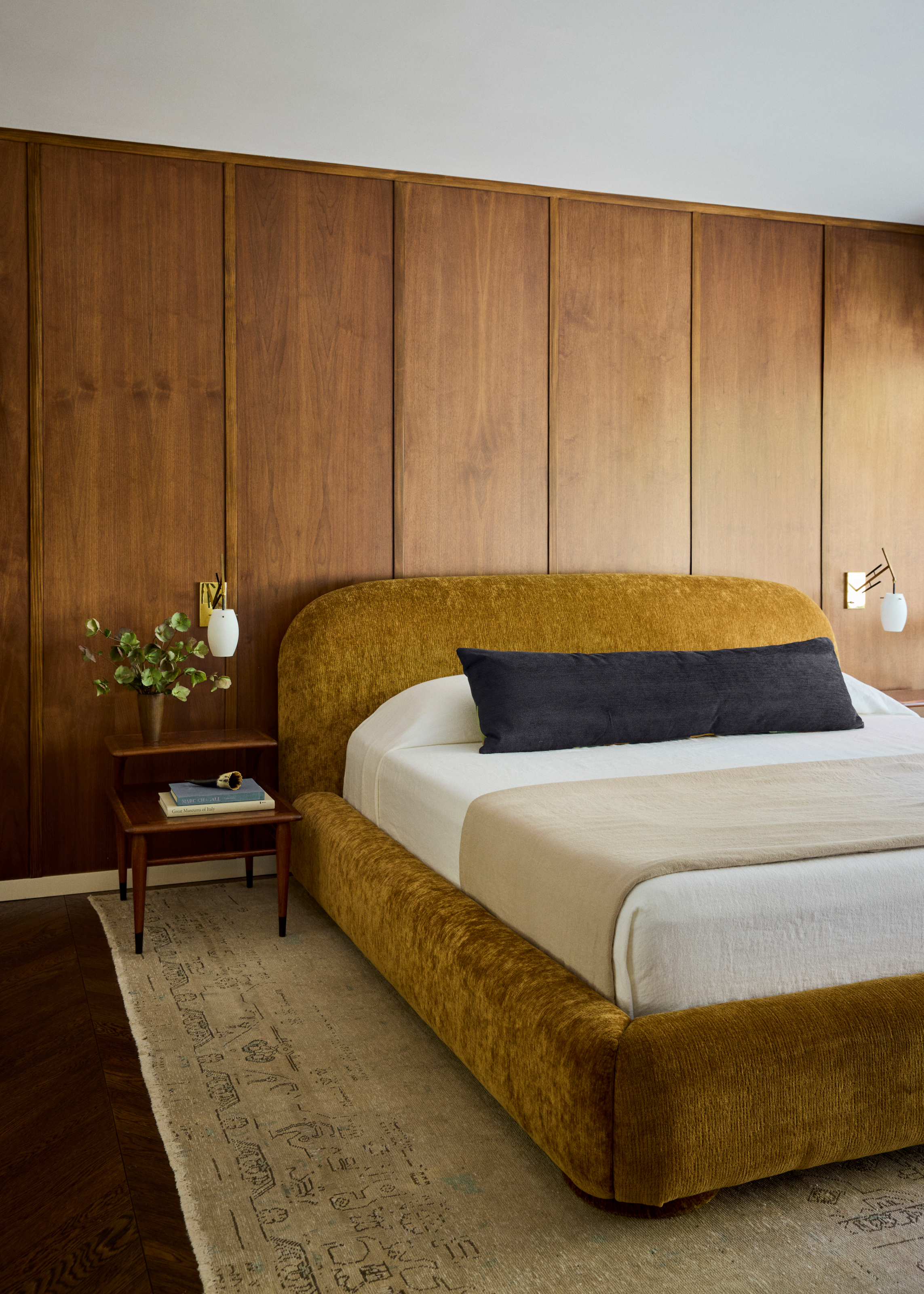 Bedroom with wood-paneled walls, ochre velvet bed, white bed linen and black accent bolster cushion