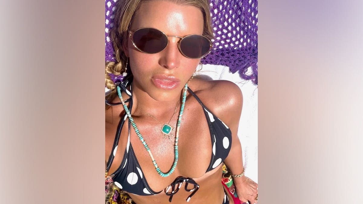 Jessica Simpson poses for a selfie while tanning in a black-and-white polka-dot bikini with layered turquoise necklaces, dangling stone earrings and dark sunglasses on her 46th birthday.