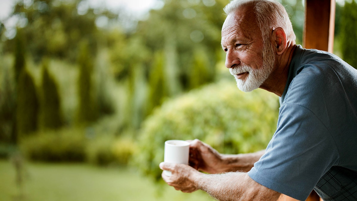 A man with white hair and a white beard holds a cup of coffee as he stands outside on a terrace.