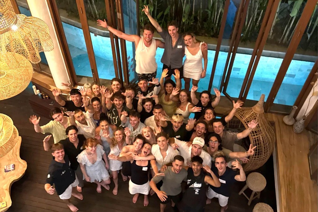 A large group of people smiling and waving towards the camera from a multi-level room with a pool.