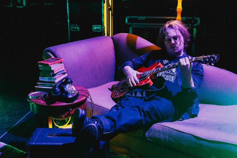 Musician playing guitar on a couch, surrounded by books and an antique telephone.