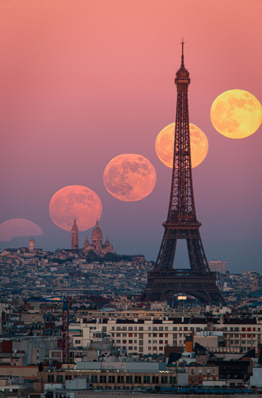 A composite image of Paris at sunset shows the Eiffel Tower and Sacré-Cœur with multiple phases of a large, orange full moon rising above the city’s skyline against a pink sky.