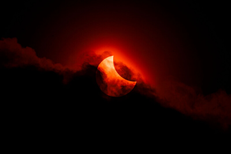 A partial solar eclipse with the sun partially obscured by the moon, glowing red and orange behind dark clouds in a dramatic night sky.
