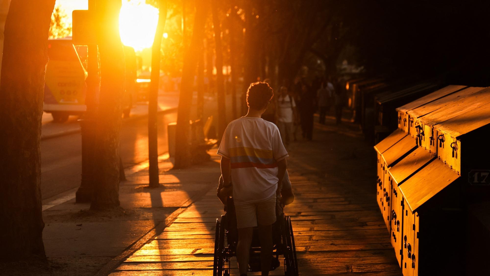 The sun beats down on a person pushing a wheelchair down the sidewalk. Taken in Paris during the 2026 heat wave.