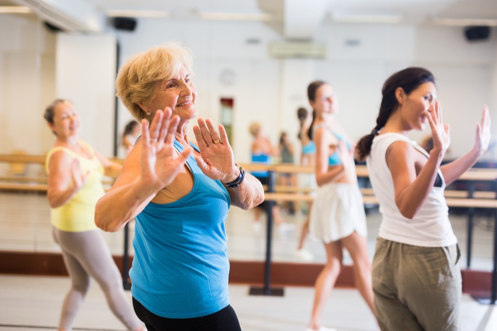 A smiling mature European woman in a blue tank top dancing energetically in a studio with other women.