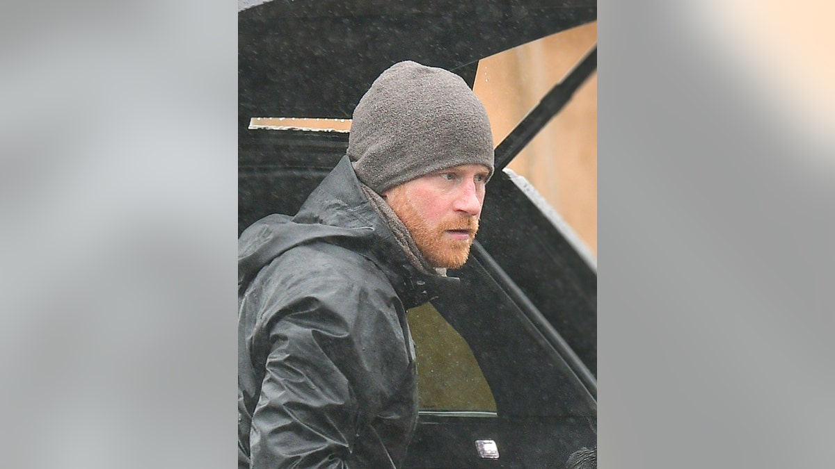 Prince Harry in a grey jacket and matching beanie standing in front of a car in Montecito looking serious.