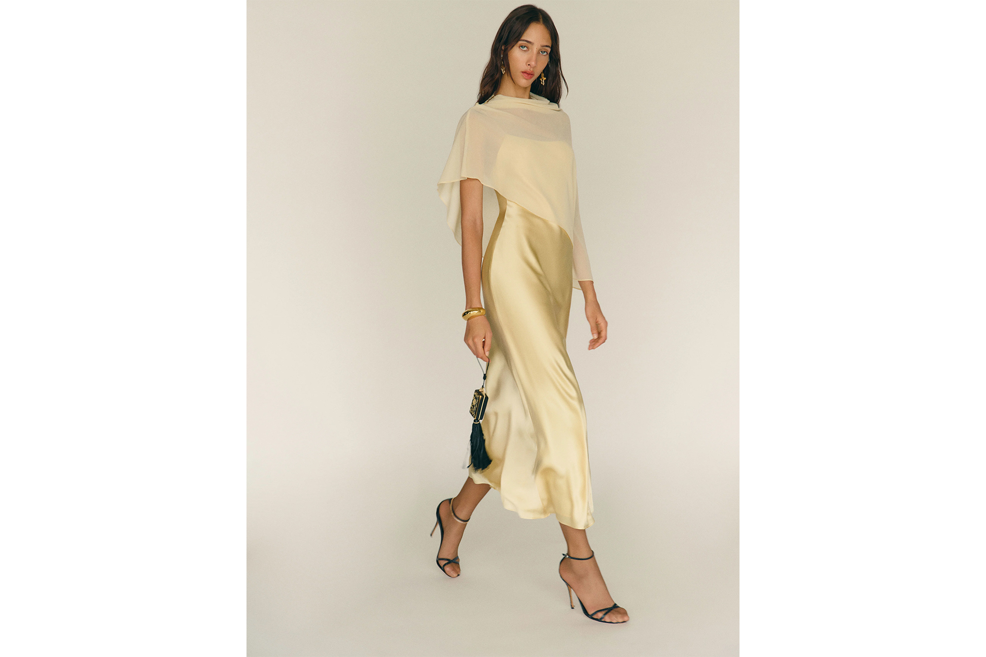 A woman in a cream-colored silk dress with a sheer draped top and black heels walking forward while holding a small black and gold purse.