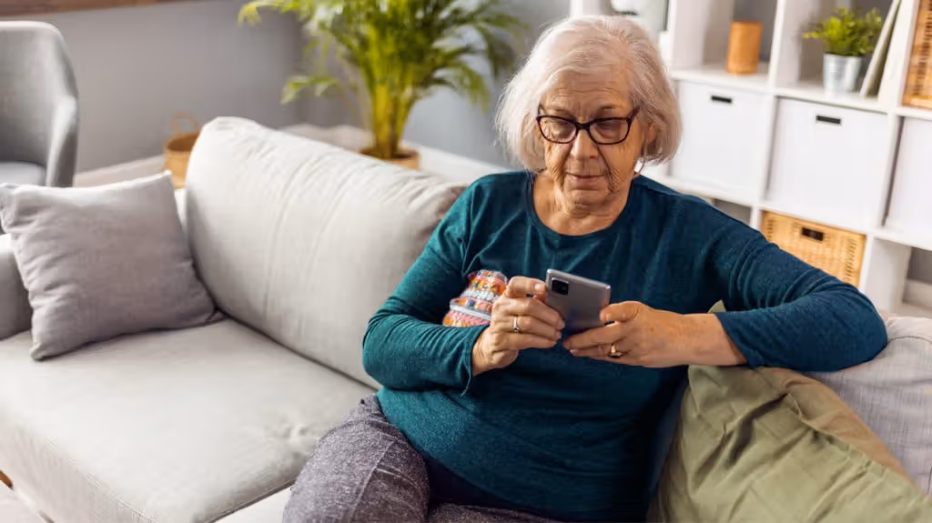 An older woman sits on a couch on her mobile phone exhibiting sedentary behavior
