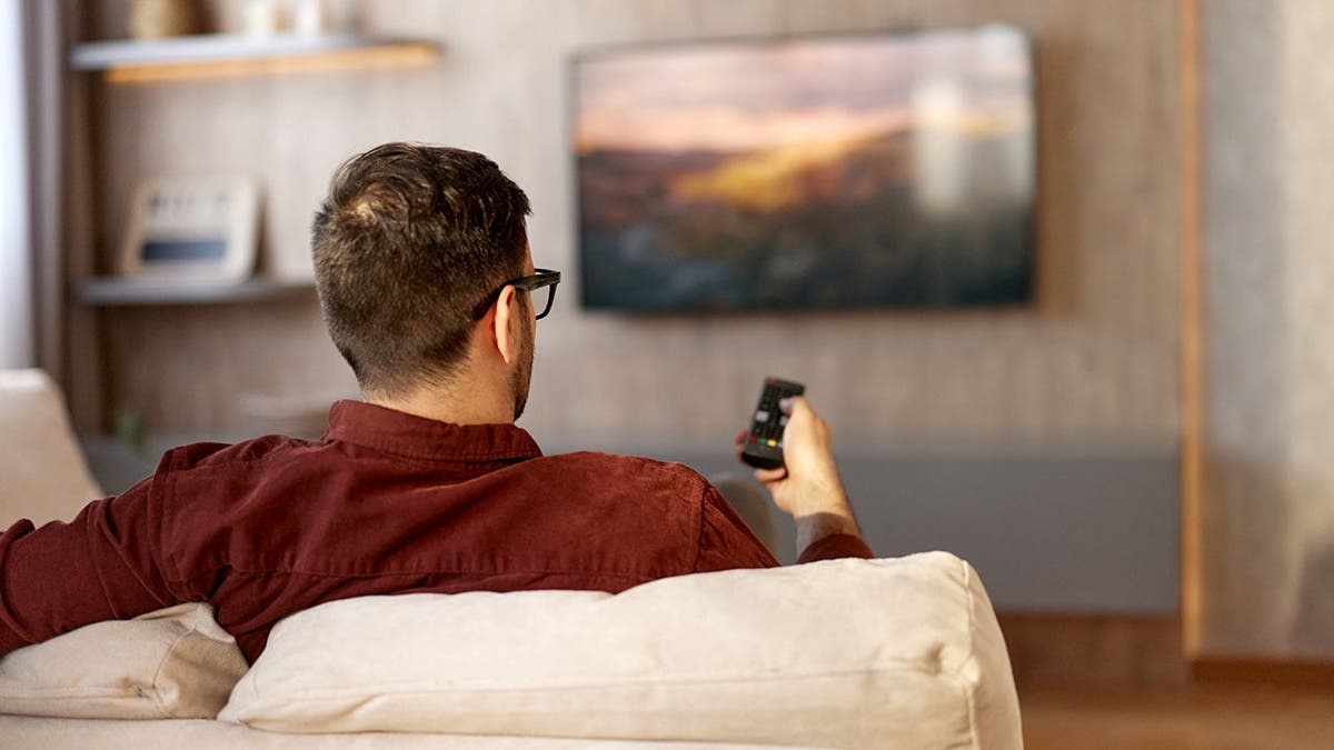 A man sits on a couch and surfs a television.