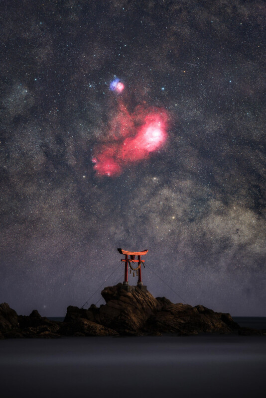 A red torii gate stands on rocky terrain by the sea under a starry night sky, with a vivid red nebula and bright stars visible above.