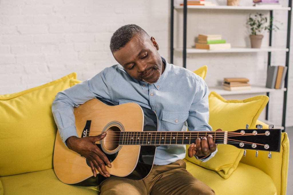 A smiling African American man playing an acoustic guitar while sitting on a yellow sofa.