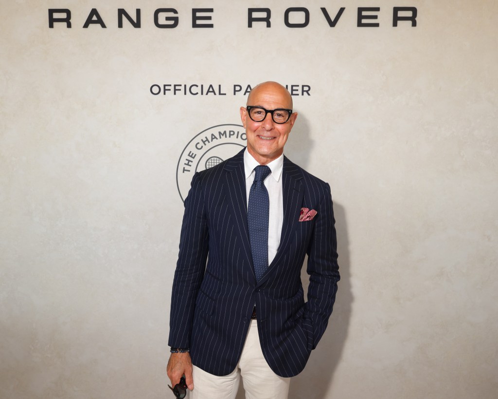 Stanley Tucci attends Wimbledon, smiling and wearing a pinstripe suit, against a backdrop with "Range Rover Official Partner" and "The Championships" logo.