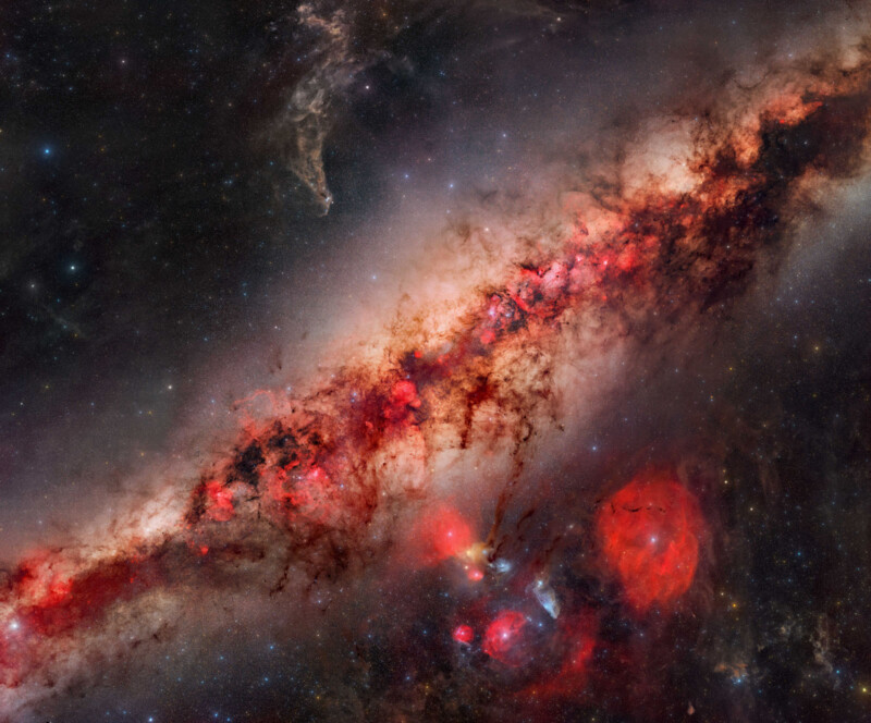 A colorful galaxy with a bright, reddish central band filled with glowing gas and dark dust lanes, surrounded by stars, nebulae, and faint cosmic clouds against a starry background.