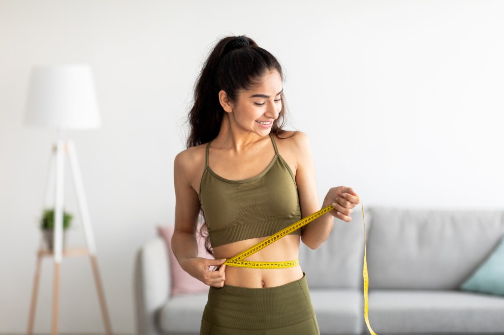 Young Indian woman in sports clothes happily measuring her waist with a yellow tape measure at home.