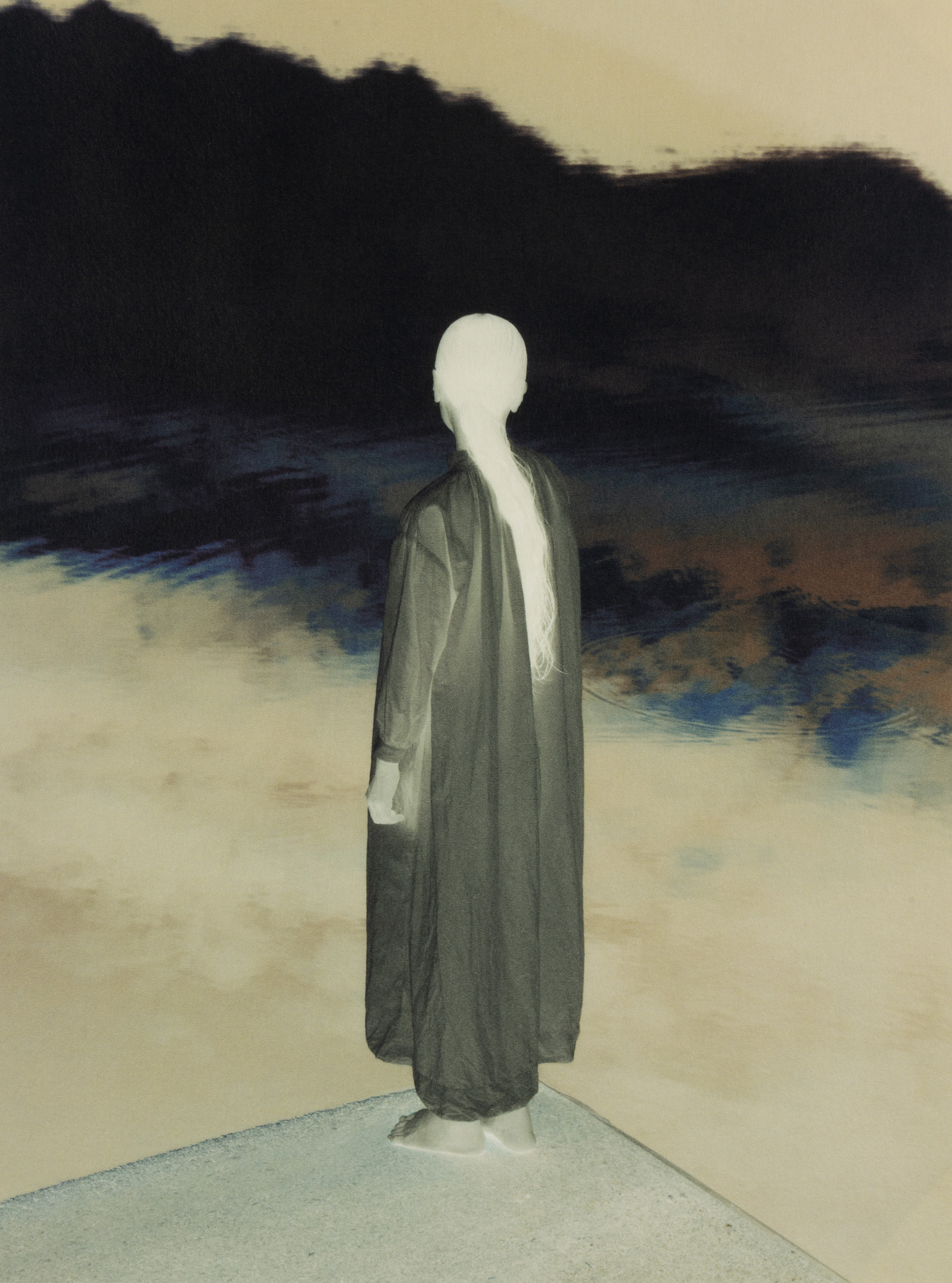Tonally inverted photograph of a pale-haired figure in a long dark robe standing on a shoreline, facing a dark landscape with a band of blue water.