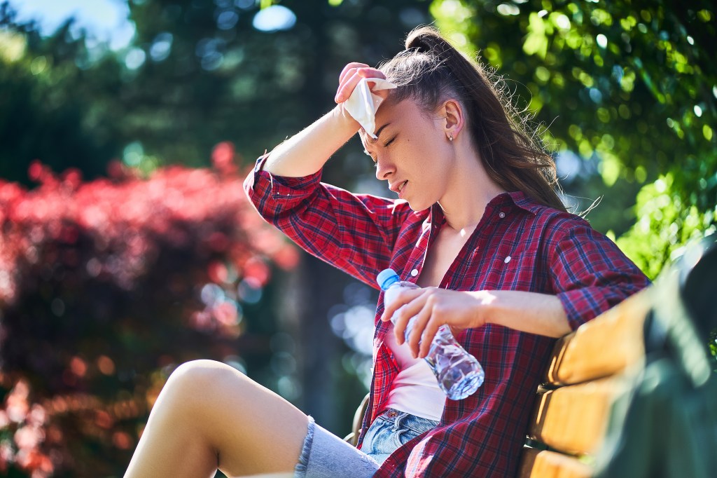 A young woman sits on a park bench, wiping sweat from her forehead with a napkin and holding a water bottle on a hot summer day.