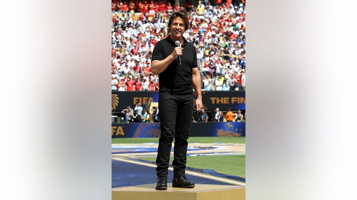 Tom Cruise speaks on the field at a soccer stadium.