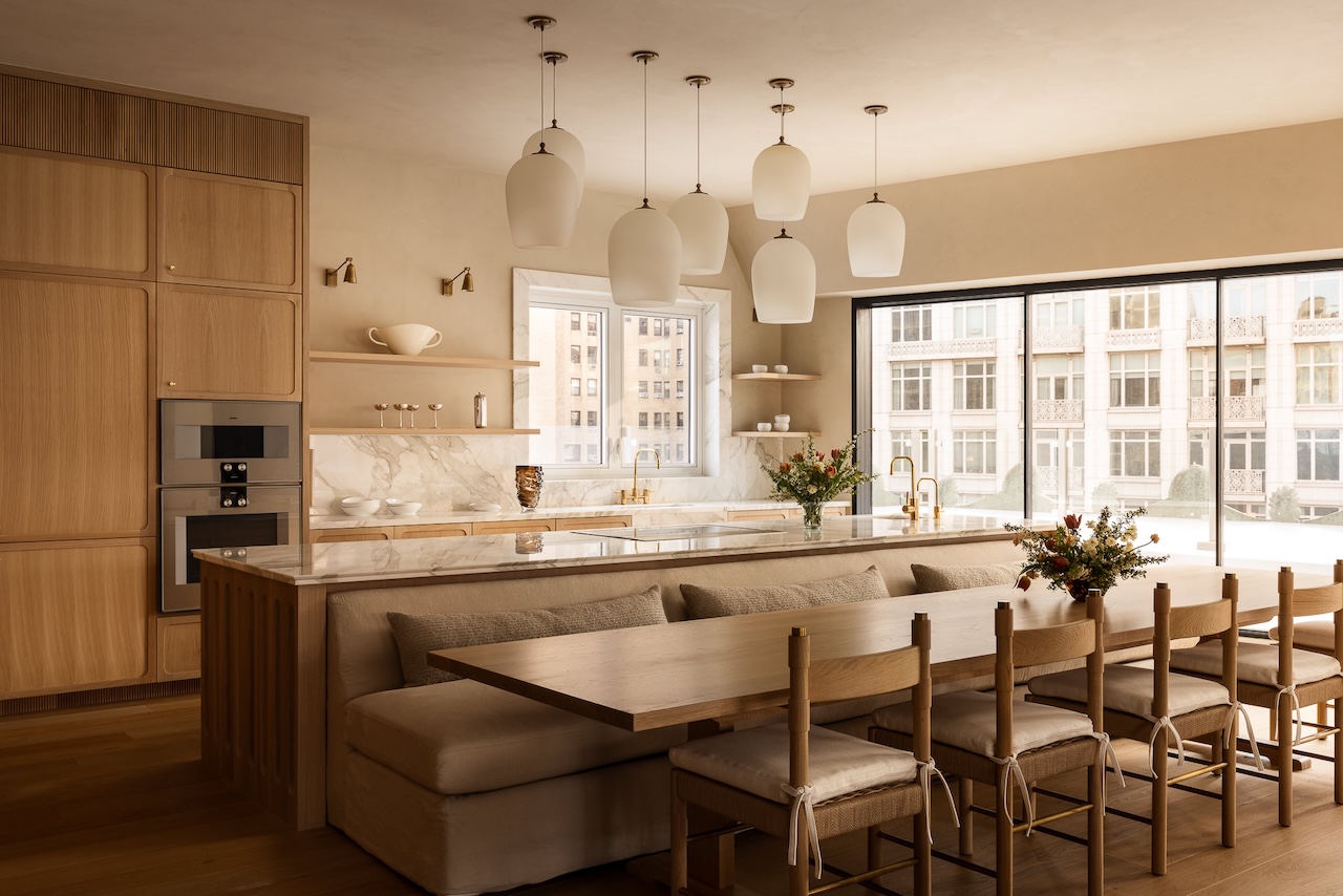 Modern kitchen with light wood cabinetry, marble countertops, beige upholstered dining bench, wooden chairs, pendant lights, and large windows overlooking city buildings.