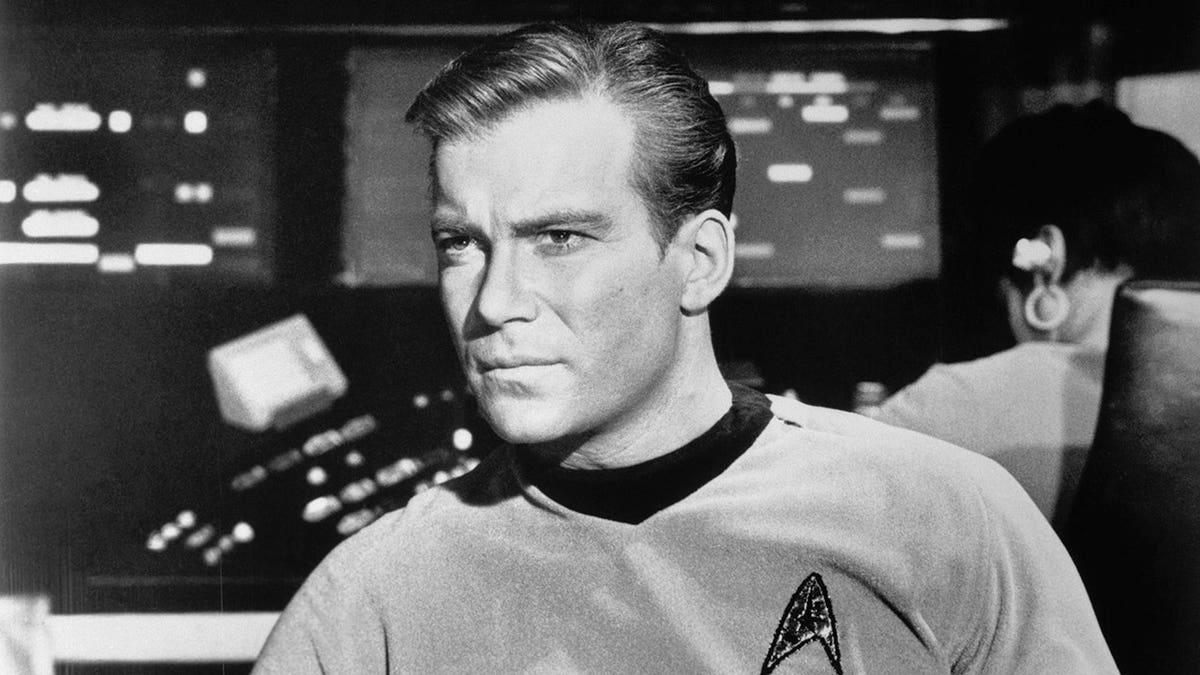 William Shatner as Captain James Kirk seated in the Starship Enterprise with a concerned expression.