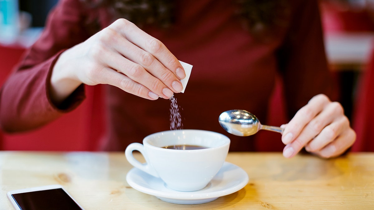 A woman adds sugar from a packet to her white cup of coffee.