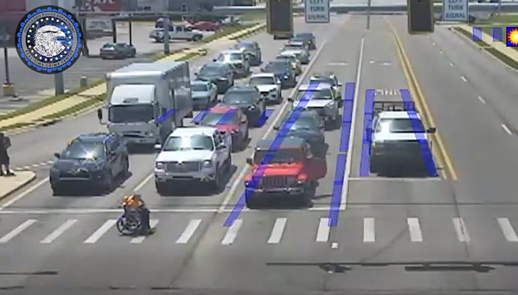 A person in a wheelchair waits at a crosswalk with heavy traffic, with a Spider-Man figure visible in the background.