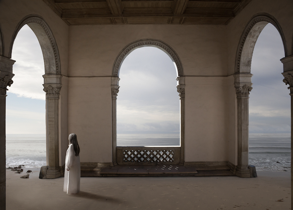 A lone figure stands beneath arched architecture facing a misty waterfront, © Ann Mitchell.