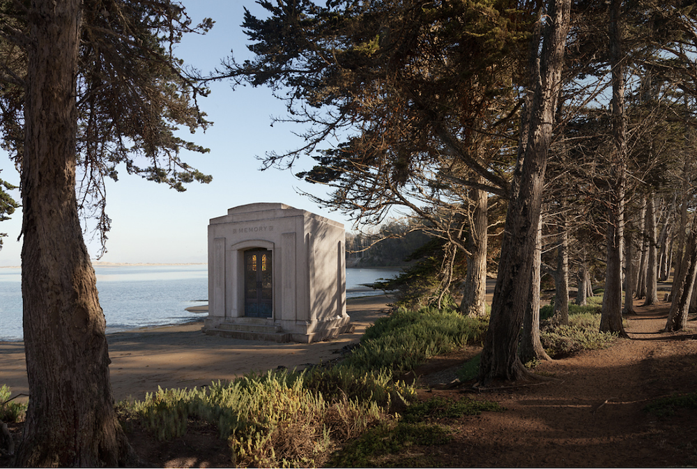 A small pale building stands among trees beside a path in warm late-day light, © Ann Mitchell.