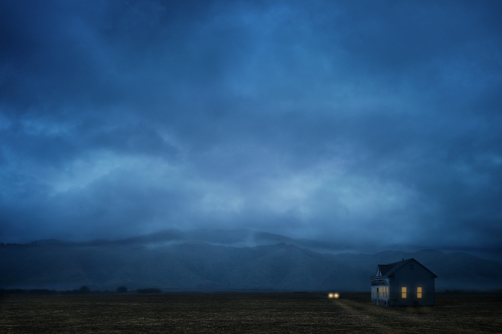 A small house sits in a dark blue landscape beneath a stormy sky, © Ann Mitchell.