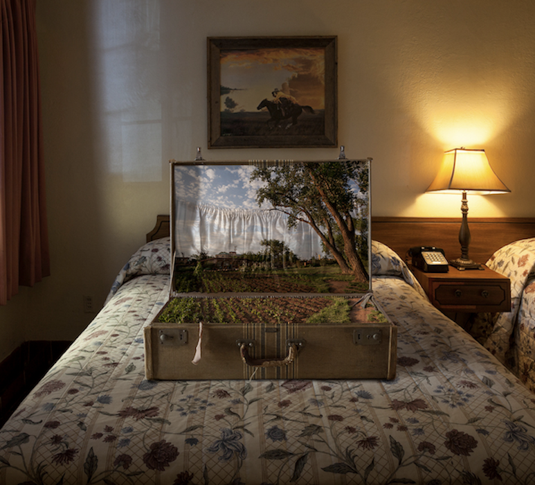 A warmly lit bedroom with a patterned bedspread and a small constructed landscape inside a suitcase placed on the bed, © Ann Mitchell.