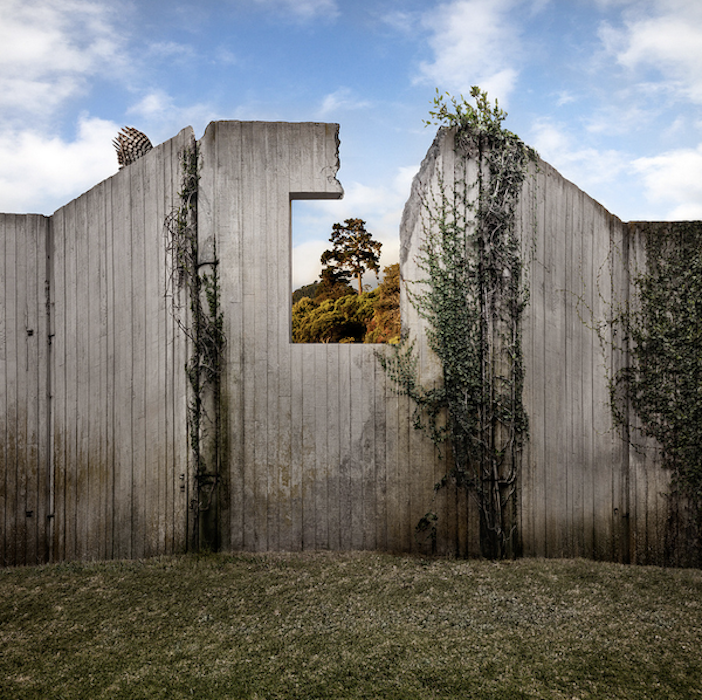 Weathered white walls and climbing vines frame a small opening onto trees and sky, © Ann Mitchell.