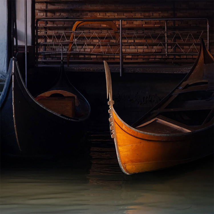 Small wooden boats rest in the deep shadows of a Venetian boathouse, one warm ochre bow catching the light above dark water. ©CynthiaGladis