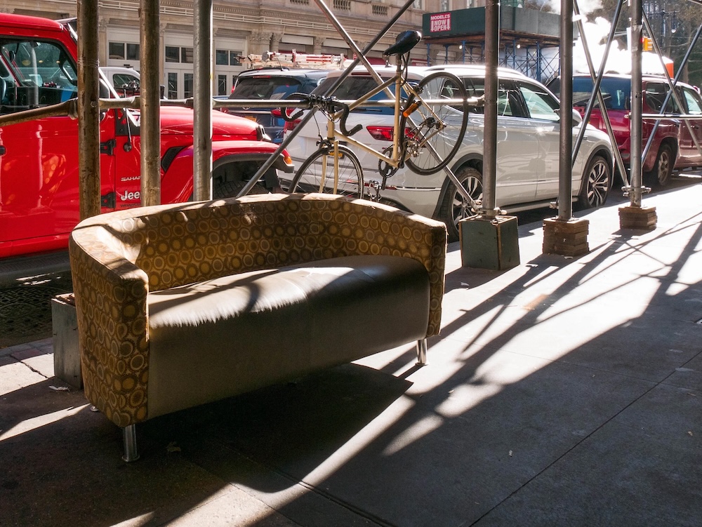 A worn patterned sofa and a bicycle share a sunlit city sidewalk beneath scaffolding, with parked cars behind them. ©CynthiaGladis