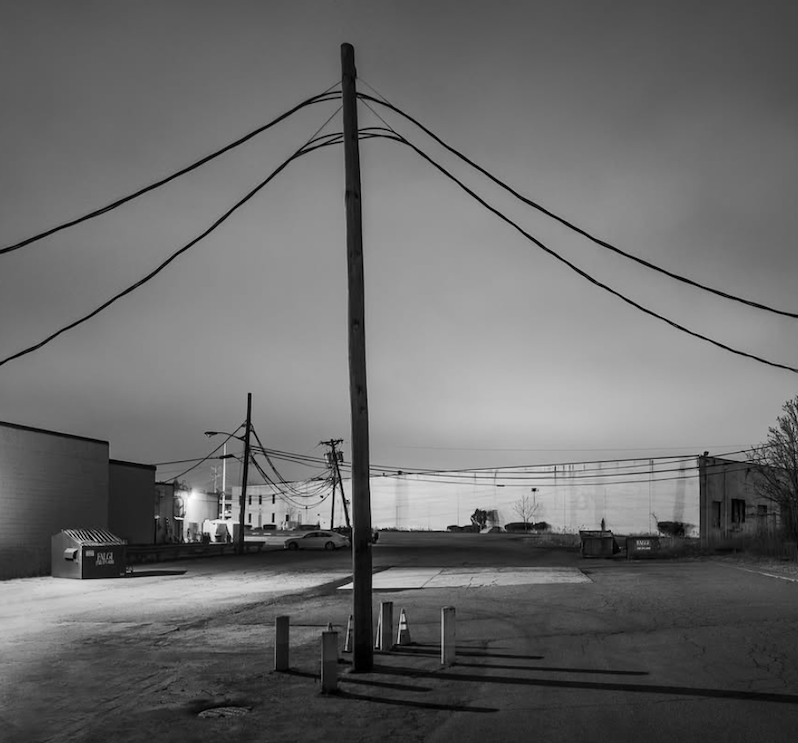Black-and-white view of a deserted industrial lot at night, crossed by utility poles and wires beneath a dark, clouded sky. ©CynthiaGladis