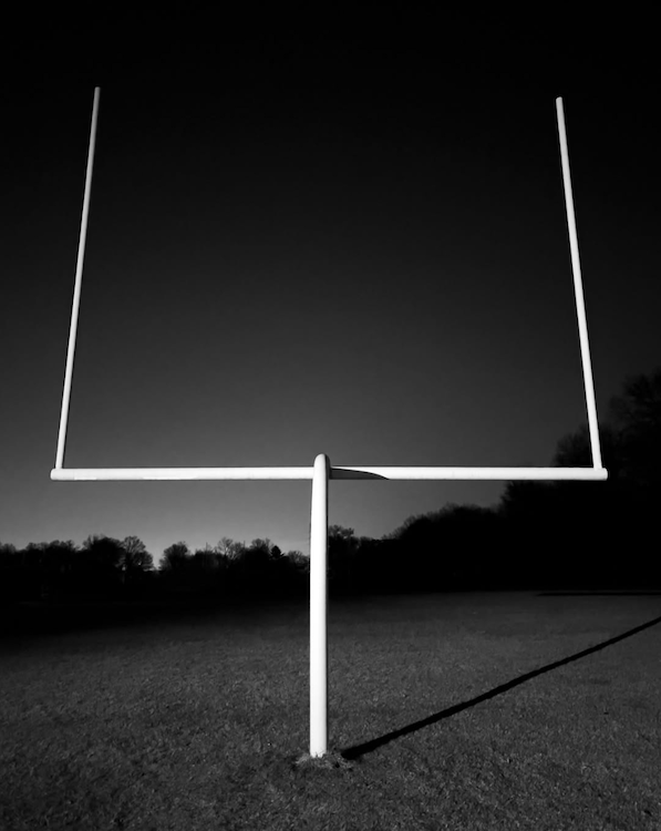 A white football goalpost rises from an empty field in a stark black-and-white scene beneath a nearly black sky. ©CynthiaGladis