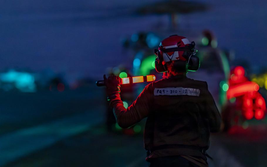 A sailor holding a lighted wand on a carrier flight deck at night. 