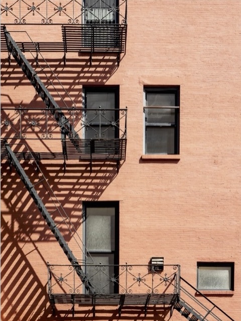 Black metal fire escapes cast angular shadows across the peach brick façade and dark windows of a city building. ©CynthiaGladis